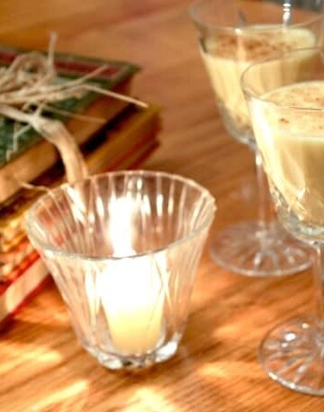 Two glasses of eggnog next to a candle and stack of old books.