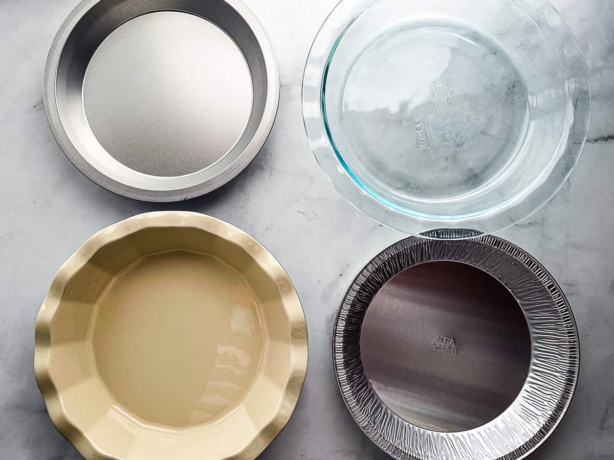 Four empty pie plates on the counter. Top row: a metal pan and a glass pan. Bottom row: ceramic pan and a disposal pan. 