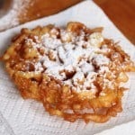 Gluten-free funnel cake dusted with powdered sugar on paper towel.