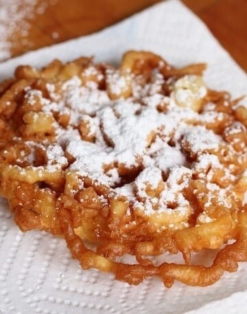 Gluten-free funnel cake dusted with powdered sugar on paper towel.