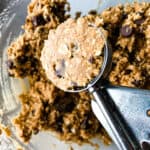 A cookie scoop filled with gluten-free oatmeal cookie dough. A bowl of dough is in the backdround.