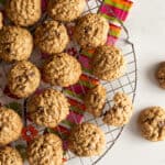 Gluten-free oatmeal cookies cooling on a wire rack. A brightly colored tea-towel sits under the wire rack. Two cookies are on the board off to the right.