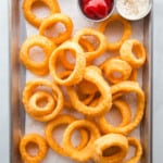 Gluten-free onion rings, along with ketchup and ranch dressing, on a baking sheet.