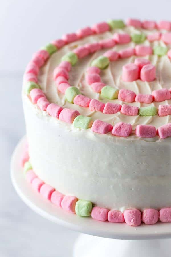 Strawberry cake with white frosting and pink and green marshmallows sits on a cake stand.