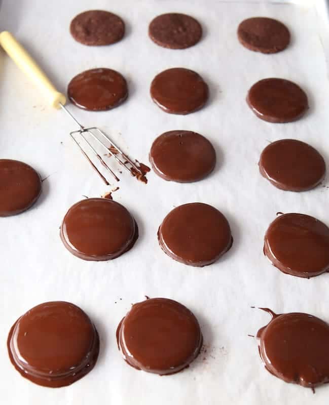 Paleo thin mints on a piece of parchment paper. Chocolate dipping tool sits on the parchment with the cookies.