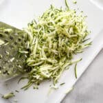 Grated zucchini on a cutting board with a box grater on the left.